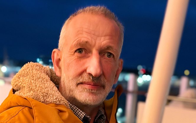 Epiphaniengemeinde Hamburg – Michael Pfeiffer – neuer Hausmeister Mann mit grauen Haaren in orangefarbener Jacke vor nächtlicher Stadtbeleuchtung.