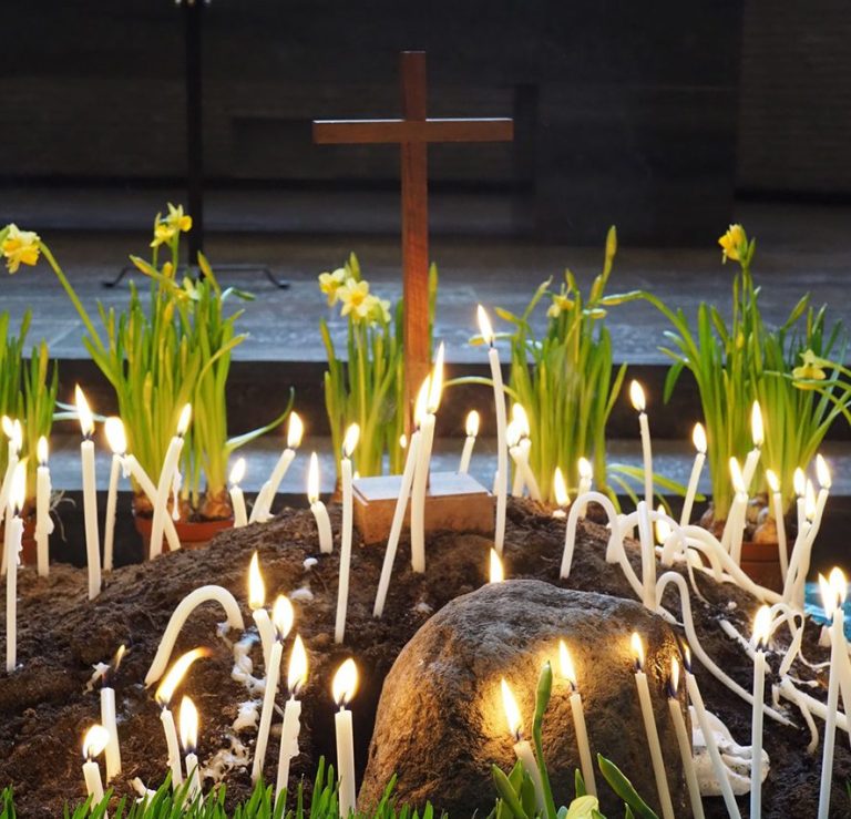 Kreuz hinter brennenden Kerzen und blühenden Narzissen auf dunklem Boden.