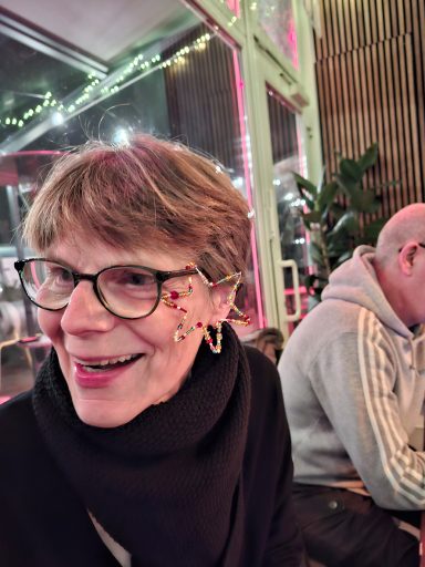 Ältere Frau mit Brille und buntem Ohrring, lächelt in einem gemütlichen Café.