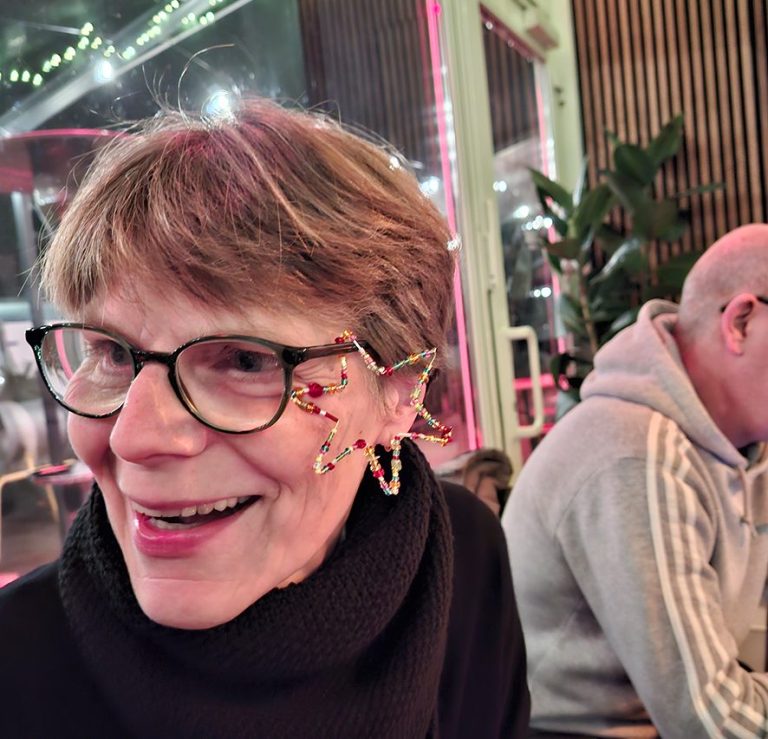 Ältere Frau mit Brille und bunten Ohrringen, lächelt vor festlicher Kulisse.