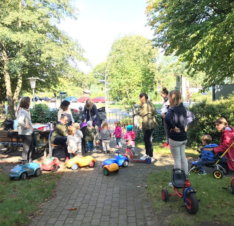 Gruppenspielplatz mit Kindern und Eltern, Spielzeugfahrzeuge auf dem Boden verteilt.