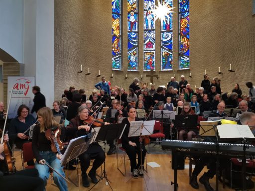 Orchesterauftritt in einer Kirche, Musiker und Sänger sitzen in Reihen, bunte Fenster im Hintergrund.