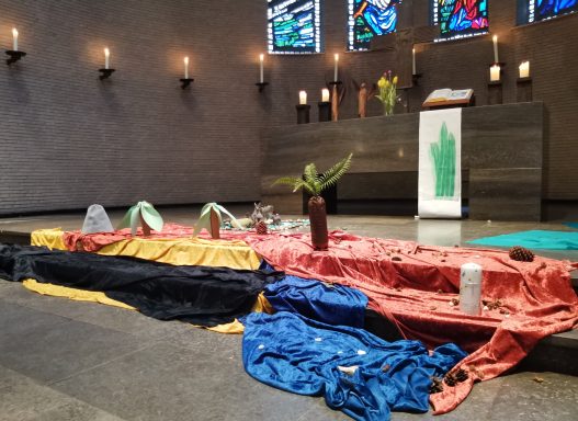 Bunte Stoffe und Pflanzen im Altarbereich einer Kirche, umgeben von Kerzen.