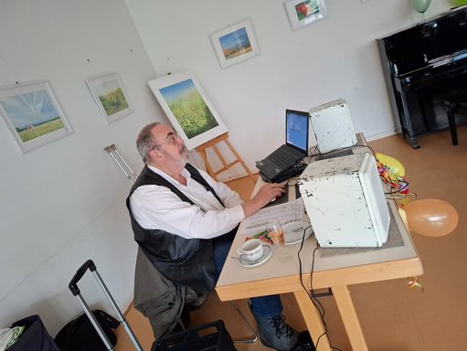 Mann sitzt am Tisch mit Laptop, umgeben von Kunst und einem Luftballon.