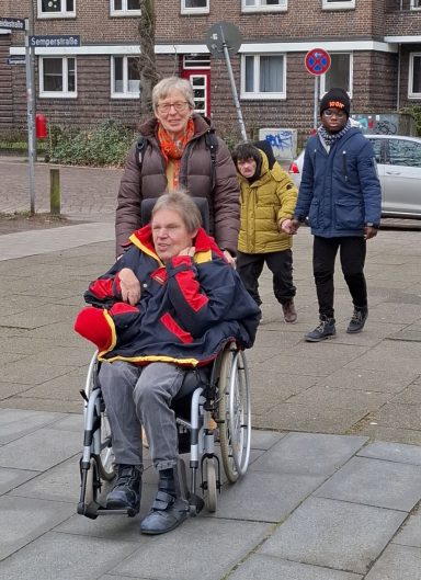 Ein Mensch im Rollstuhl wird von einer  Frau geschoben und von zwei weiteren Menschen begleitet, die sich an den Händen fassen.