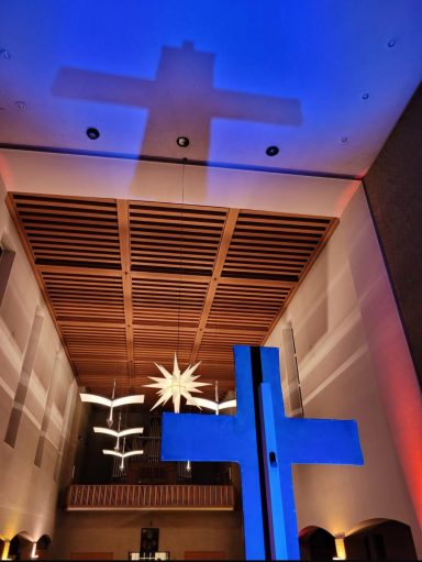 Großes Kreuz im Kirchenraum mit blauen und roten Lichtakzenten und Deckenbeleuchtung.