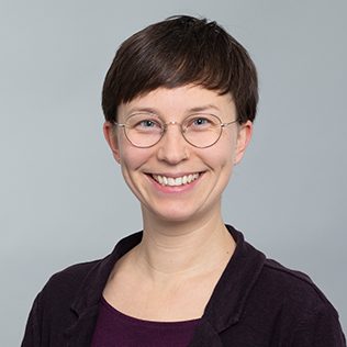 Pastorin Friederike Arnold mit kurzen Haaren, Brille und lächelndem Gesicht, trägt ein dunkles Shirt und einen dunklen Blazer.