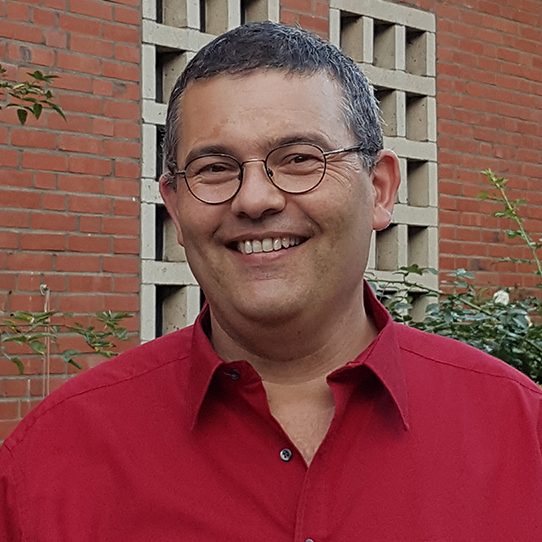 Broder Jürgensen, Vorsitzender des Kirchengemeinderats, lächelnd mit Brille in rotem Hemd vor Rosensträuchern und Backsteinwand der Kirche.