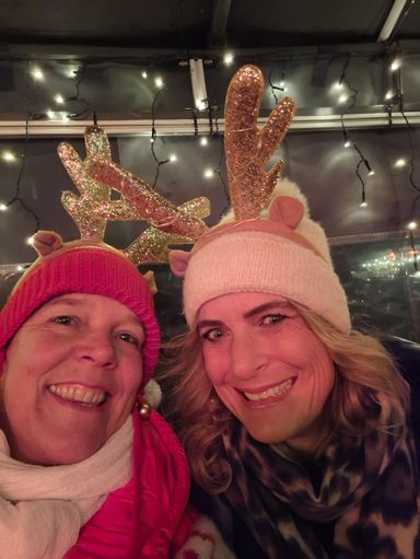 Zwei Frauen mit Weihnachtsmützen und Rentiergeweih, lächelnd in festlicher Umgebung.