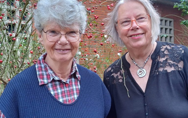 Zwei lächelnde Frauen, Jette Wrobel und Heike Schoof, stehen vor blühenden Rosen vor der Epiphanienkirche.