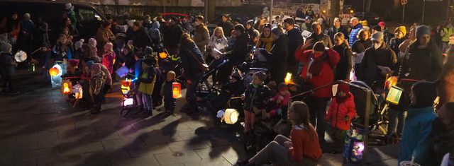 Große Gruppe von Kindern und Erwachsenen in der Dunkelheit mit leuchtenden Laternen auf dem Kirchenvorplatz.