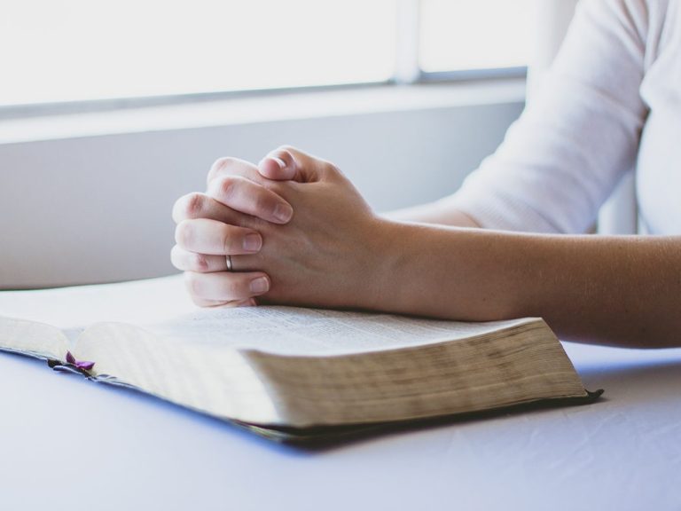 Hände falten sich zum Gebet über einer offenen Bibel auf einem Tisch.