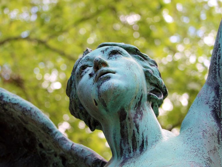Grüne antike Kupferstatue eines Engels mit erhobenen Armen, Blick nach oben unter Bäumen.
