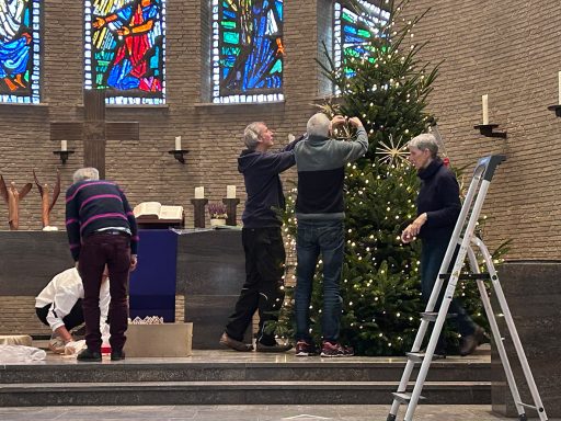 Vier Personen dekorieren einen Weihnachtsbaum in einer Kirche. Im Hintergrund sind Fenster mit Glasmalerei.